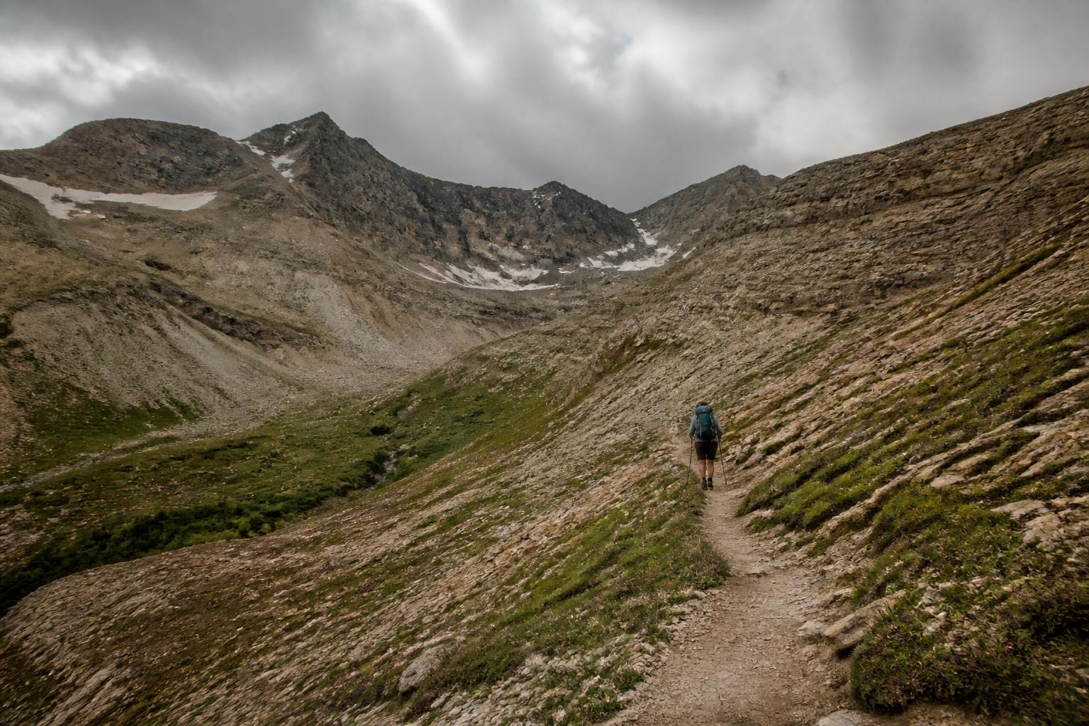 A single hiker ascending a narrow alpine trail toward a high mountain pass